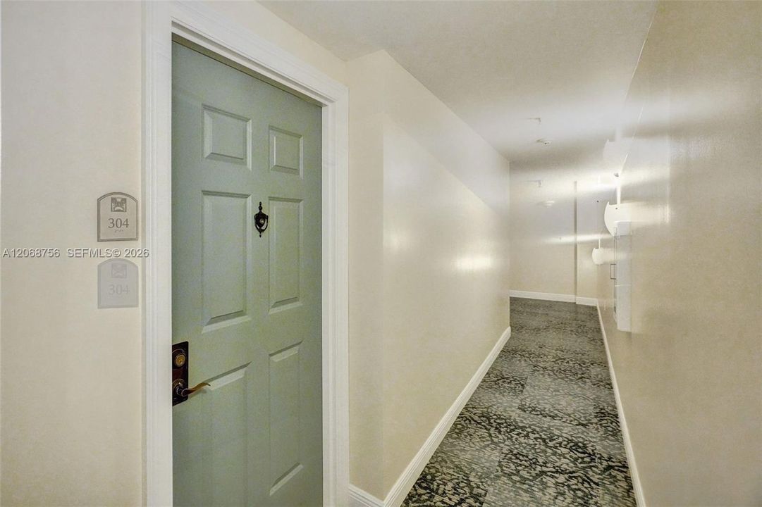 Paneled door with a decorative knocker, lever-style handle, and wall-mounted unit identifier, set within a corridor featuring patterned carpeting