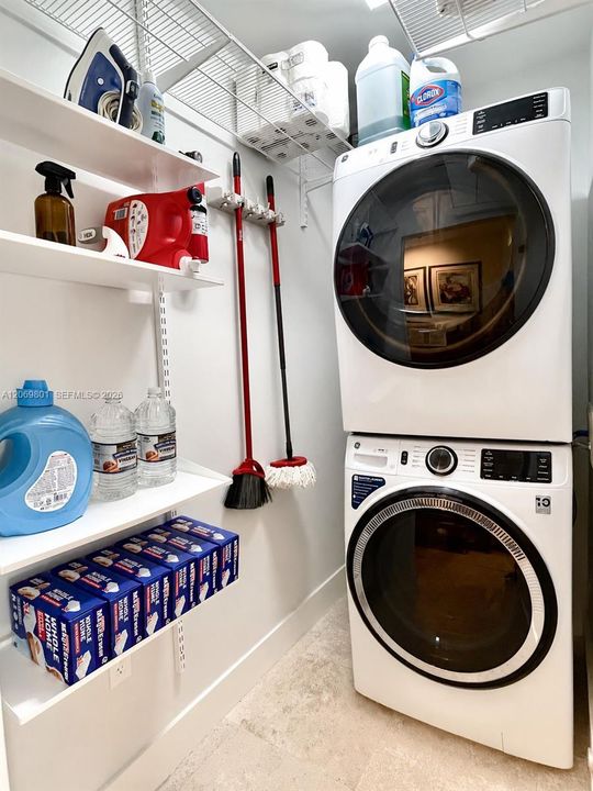 Bright and functional laundry room featuring a stacked washer and dryer, extensive open shelving, overhead wire storage, and dedicated space for cleaning supplies and household essentials. The organized layout maximizes storage while keeping everything easily accessible in a clean, efficient setting.