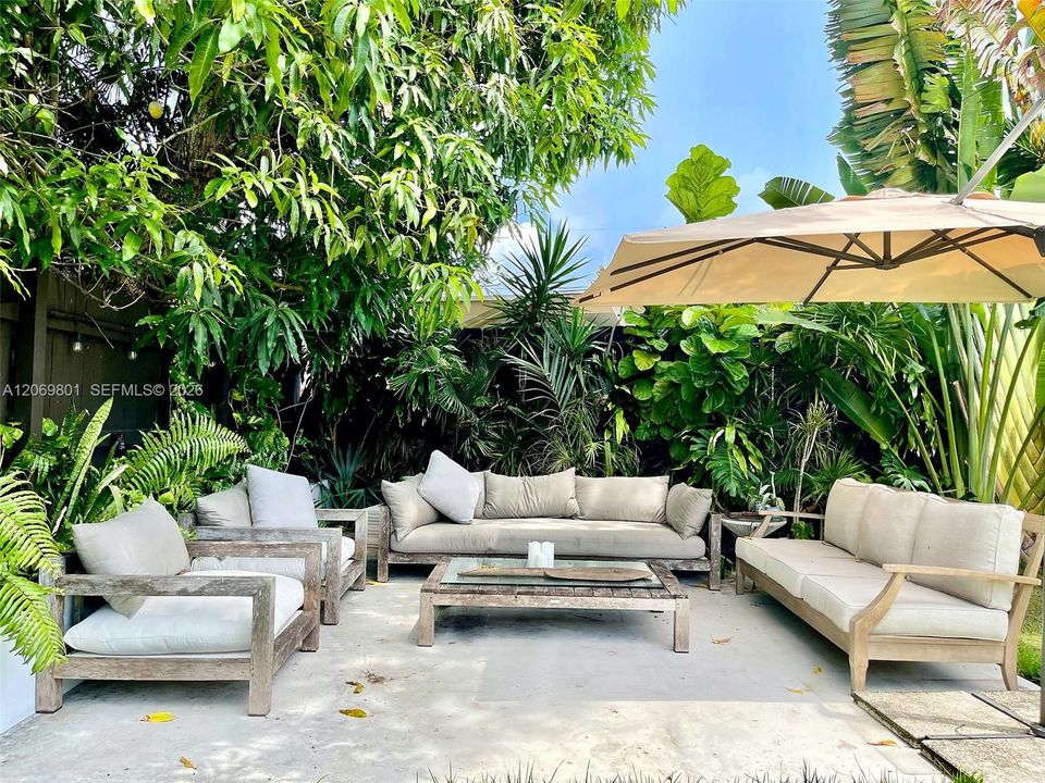 Inviting covered patio seating area surrounded by lush tropical landscaping. The spacious arrangement includes multiple sofas and lounge chairs, a central coffee table, and a large umbrella for added shade, creating a private and comfortable setting for relaxing, entertaining, or enjoying the outdoors.