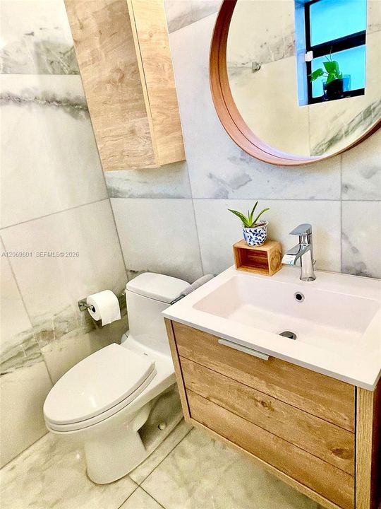 Stylish half bathroom featuring marble-style finishes, a floating wood vanity with a modern rectangular sink, coordinating wall-mounted storage, and a round wood-framed mirror. Natural light, clean lines, and warm wood accents create a bright and welcoming space for guests