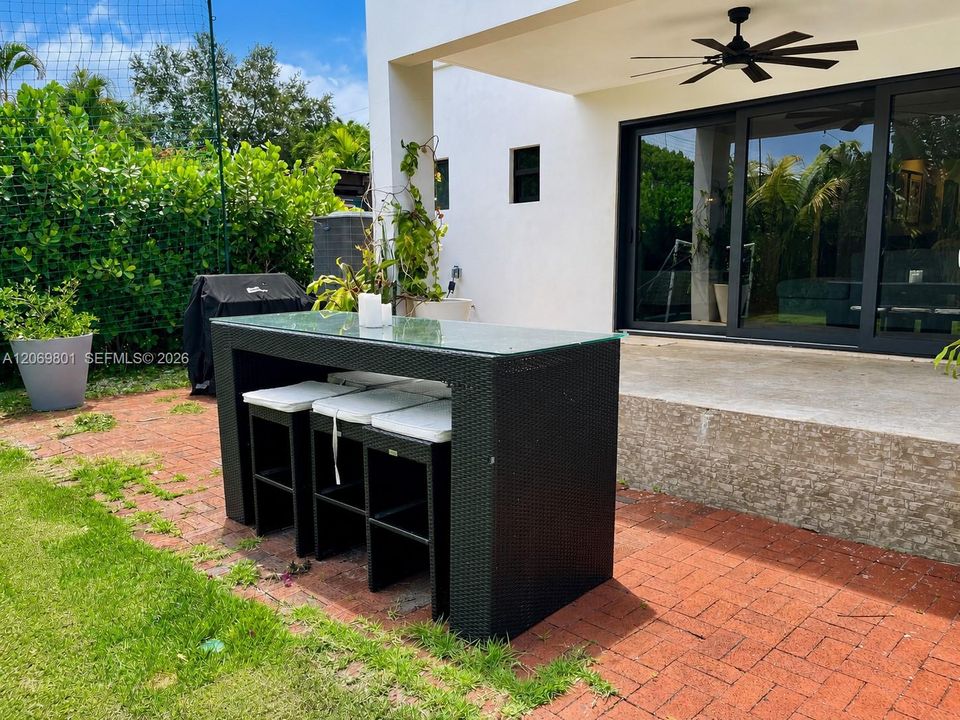 Versatile outdoor entertaining area featuring a covered patio with a ceiling fan, large sliding glass doors, and a bar-height dining table with seating. The adjoining brick patio includes space for grilling and connects directly to the lawn, creating a practical setting for outdoor meals, gatherings, and everyday relaxation.