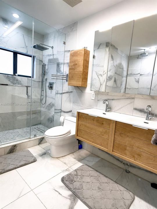 Bright and modern master bathroom featuring marble-style finishes, a glass-enclosed walk-in shower with rainfall and handheld fixtures, a double-sink vanity with wood cabinetry, mirrored storage, and a sleek contemporary toilet. Recessed and vanity lighting enhance the clean, polished design.