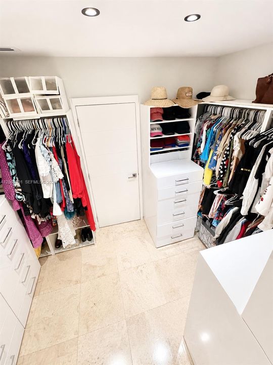 Spacious walk-in closet featuring built-in cabinetry, abundant drawers, open shelving, and extensive hanging space on both sides. Recessed lighting and light stone flooring create a bright, organized setting with generous room for clothing, shoes, hats, handbags, and accessories.