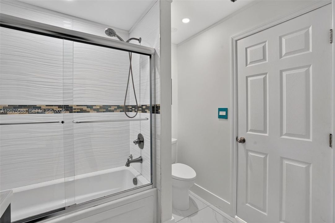 Bathtub and shower combination featuring textured horizontal wall tile, an inlaid decorative mosaic tile border, a glass sliding door enclosure, and a metallic finish rainfall showerhead