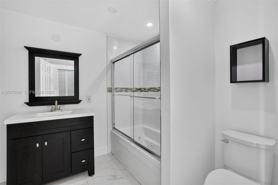 Bathroom featuring a dark-finish vanity with an integrated basin, a black-framed wall mirror, a glass-enclosed tub with tiled surround, and a wall-mounted shelving unit