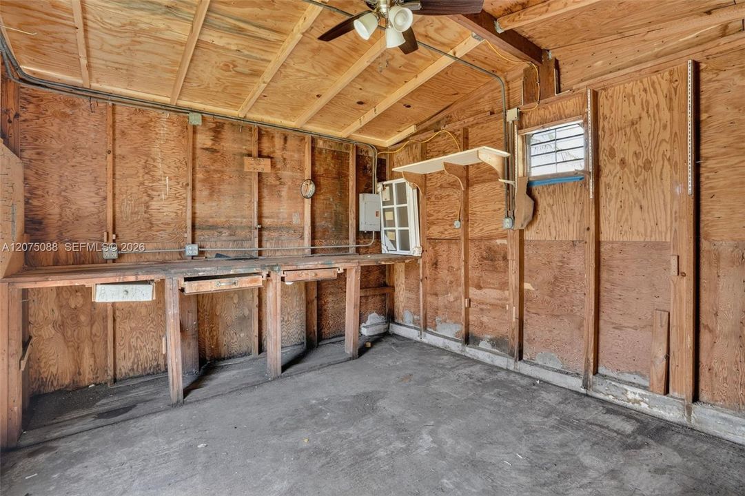 Workspace area featuring plywood wall paneling, a built-in wooden workbench with drawers, a ceiling-mounted fan, and a grid-pane window