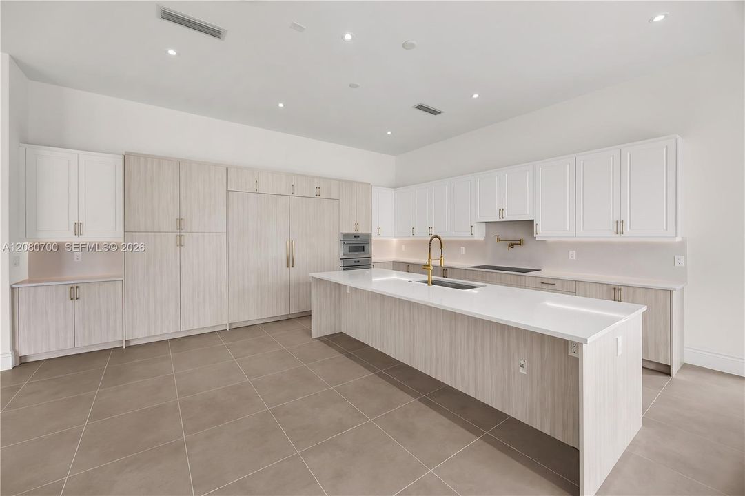 Expansive kitchen featuring a large central island with a sink, extensive white cabinetry, light wood-finish cabinetry, built-in stainless steel appliances, and sleek tile flooring