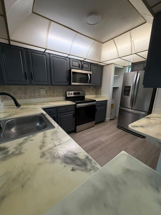 Kitchen featuring dark-toned cabinetry, stainless steel appliances, patterned countertops, wood-finish flooring, and a tray ceiling with recessed lighting