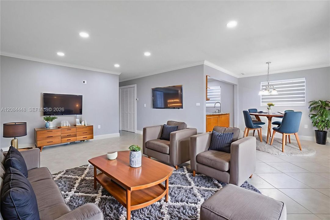 Step into an airy and open living space filled with natural light and timeless charm. The living room features neutral tile flooring, soft lavender-gray walls, and elegant crown molding for a polished finish. Recessed LED lighting enhances the fresh and modern ambiance