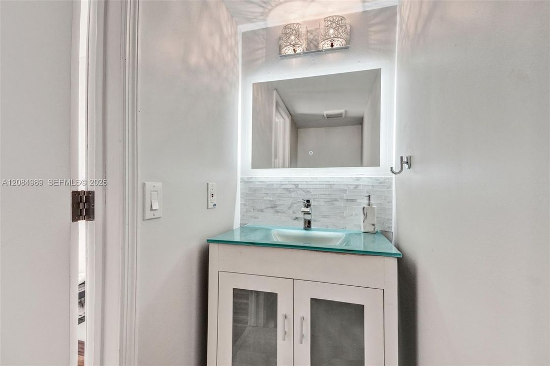 Vanity featuring a glass countertop with an integrated basin, white cabinetry with frosted glass inserts, LED-backlit mirror, and a textured tile backsplash