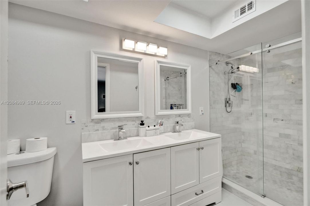 Double vanity with rectangular undermount sinks, stone-finish countertops, framed mirrors, glass-enclosed shower with tiled surround, and recessed ceiling lighting