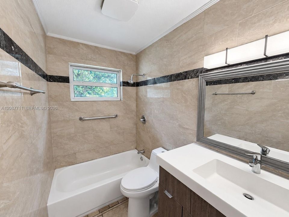 Full-height neutral stone tiling with decorative dark stone accent banding, modern wood-finish vanity with integrated rectangular sink, wall-mounted multi-light fixture, and built-in grab bars