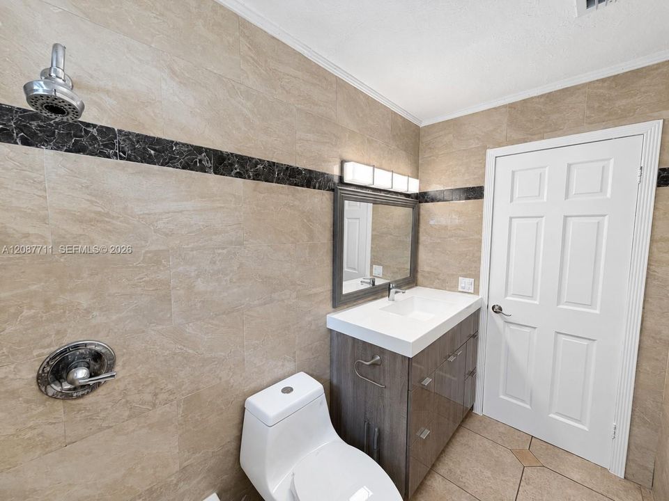 Full-wall tile surround with dark stone accent banding, wood-finish vanity with integrated sink, four-light vanity fixture, and six-panel interior door
