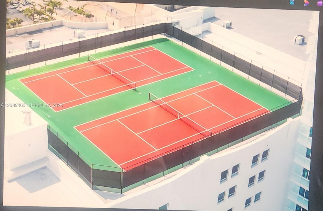 Rooftop amenity featuring two full-size tennis courts with red colored playing surfaces and green surrounds, enclosed by a perimeter fence