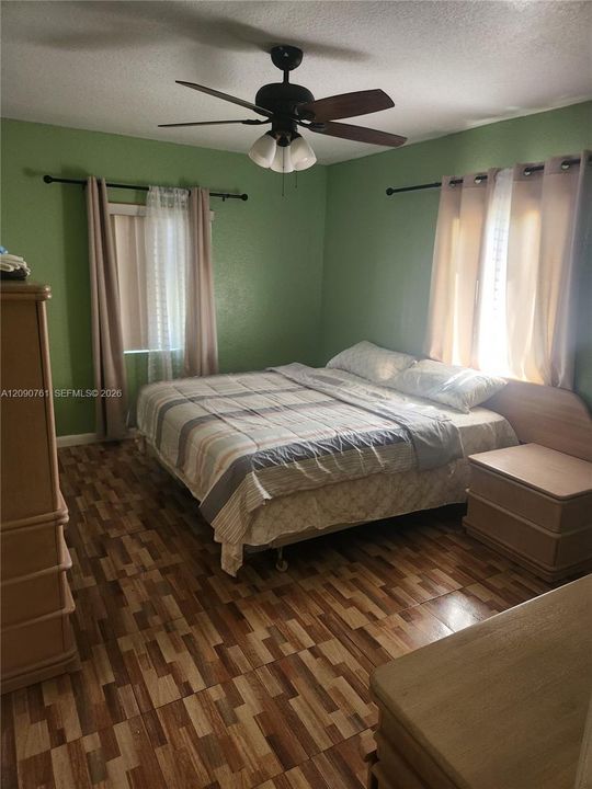 Multi-tonal wood-finish flooring, two large windows with black curtain rods, and a ceiling-mounted fan with integrated lighting