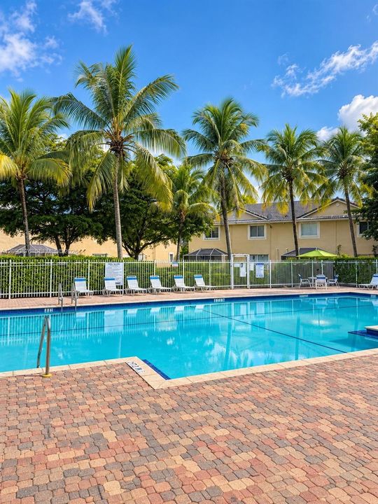 Outdoor swimming pool with a paved brick deck, surrounded by palm trees and a perimeter fence