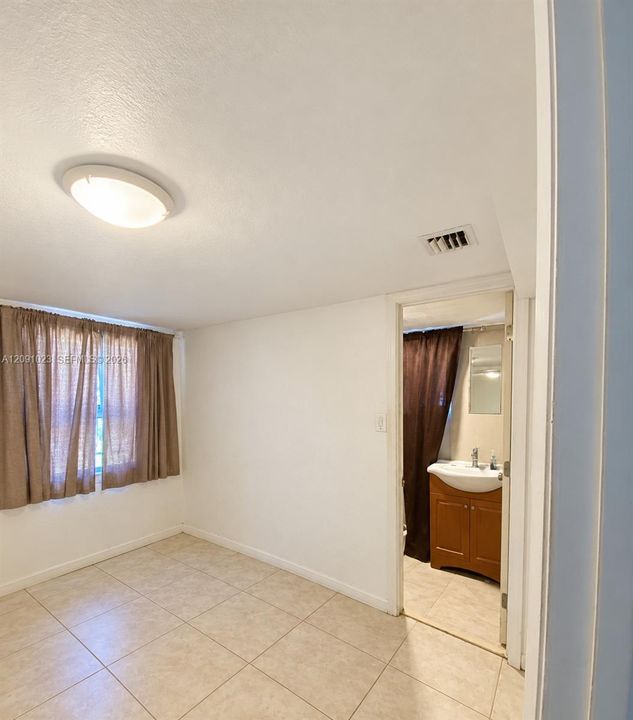 Room featuring tile flooring and a flush-mount ceiling light fixture, opening into a bathroom equipped with a wood-finish vanity, integrated sink, and wall-mounted mirror