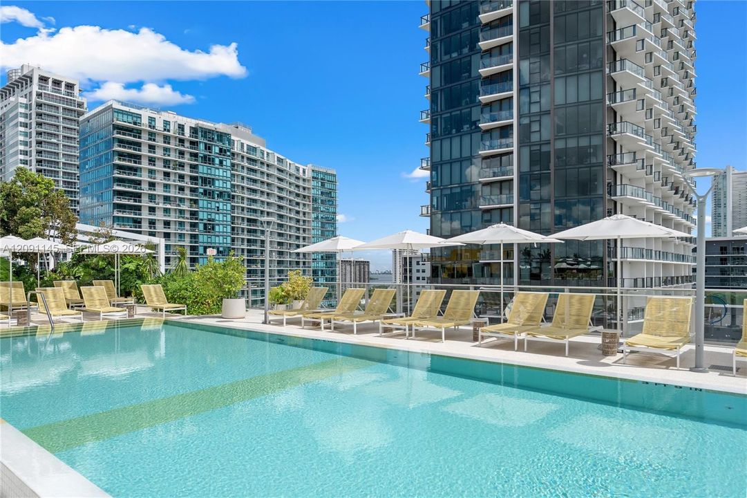 Resort-style pool deck featuring a swimming pool, lounge chairs, and sun umbrellas, surrounded by high-rise buildings