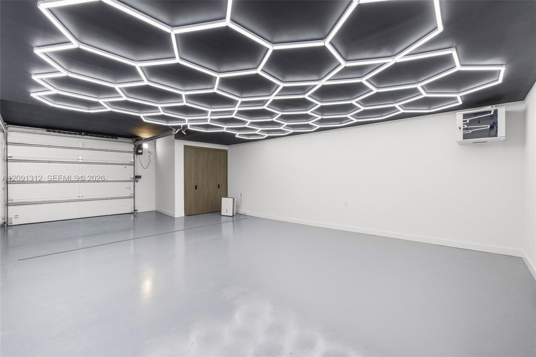 Custom ceiling with hexagonal LED light panels, smooth-finish gray flooring, white overhead garage door, and flat-panel wood-finish storage cabinetry