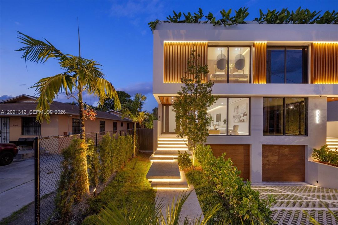 Modern architectural exterior featuring a stucco facade, vertical wood-slat accents, floor-to-ceiling glass windows, integrated staircase lighting, and a permeable paver driveway