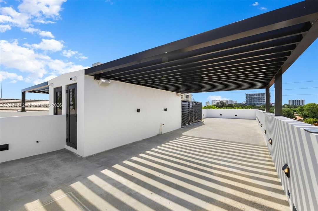 Rooftop terrace featuring a flat concrete surface, a black metal pergola, wall-mounted exterior lighting, and solid perimeter walls