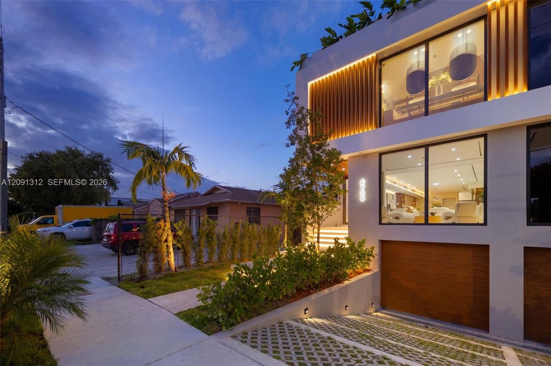 Contemporary multi-story exterior featuring floor-to-ceiling glass windows, integrated accent lighting, wood-slat architectural detailing, and a multi-car subterranean-level garage with a concrete paver driveway