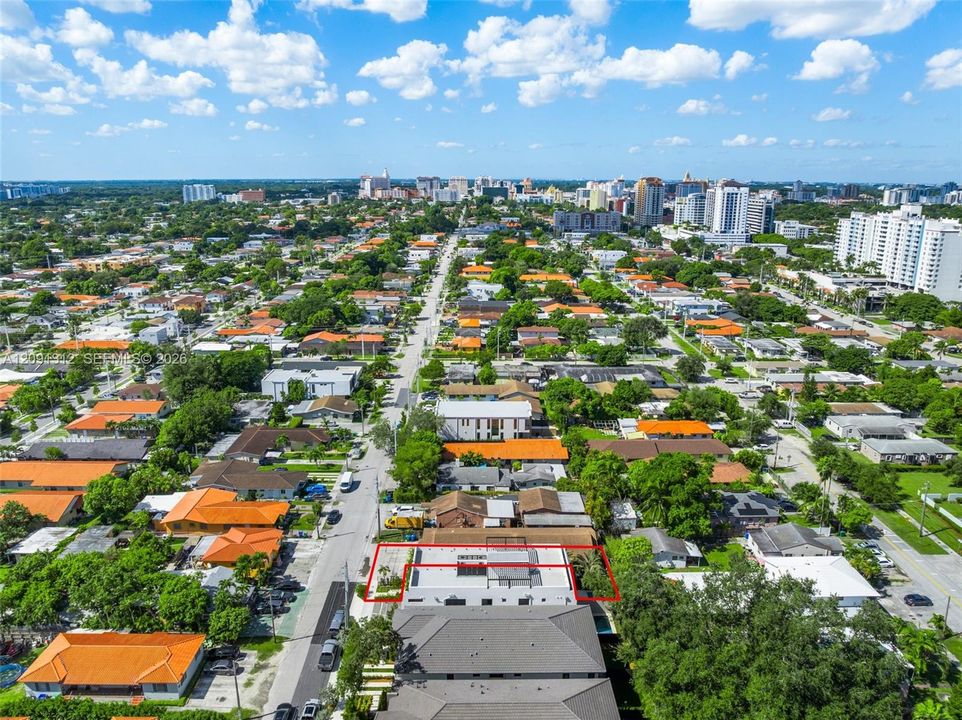 Aerial perspective of a multi-story property featuring a flat roofline, integrated rooftop terrace, ground-level paved surfacing, and mature perimeter landscaping within a dense urban landscape