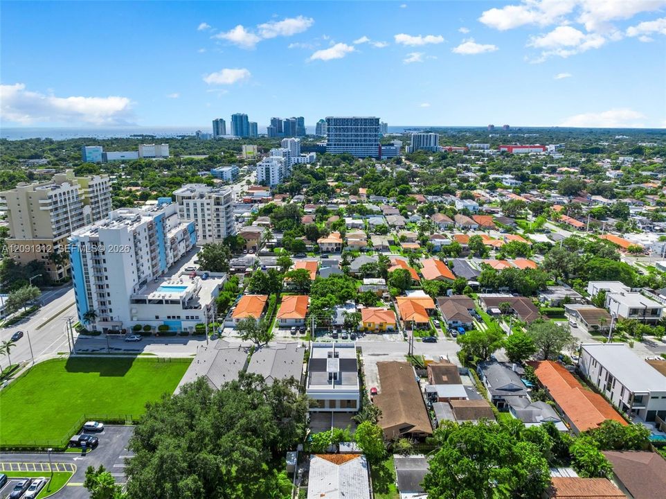 Aerial perspective of a dense urban environment featuring multi-story residential buildings, paved thoroughfares, manicured green spaces, and mature tree canopies