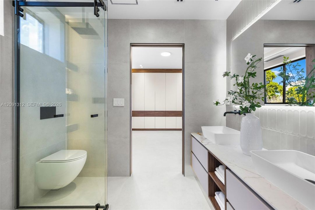 Bathroom featuring a glass-enclosed shower with matte black hardware, wall-mounted commode, and dual-sink vanity with light-toned countertop and open shelving