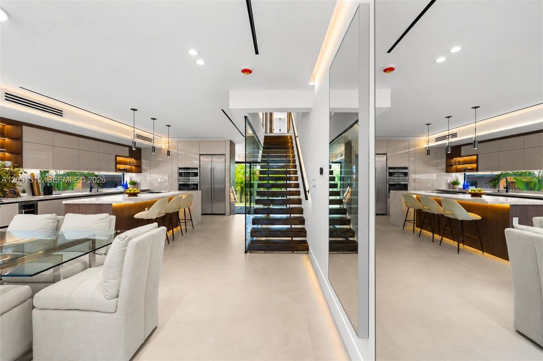 Modern open-concept area featuring sleek flat-panel cabinetry, a central island with integrated lighting, polished stone countertops, and a staircase with wood-tread risers and glass balustrades