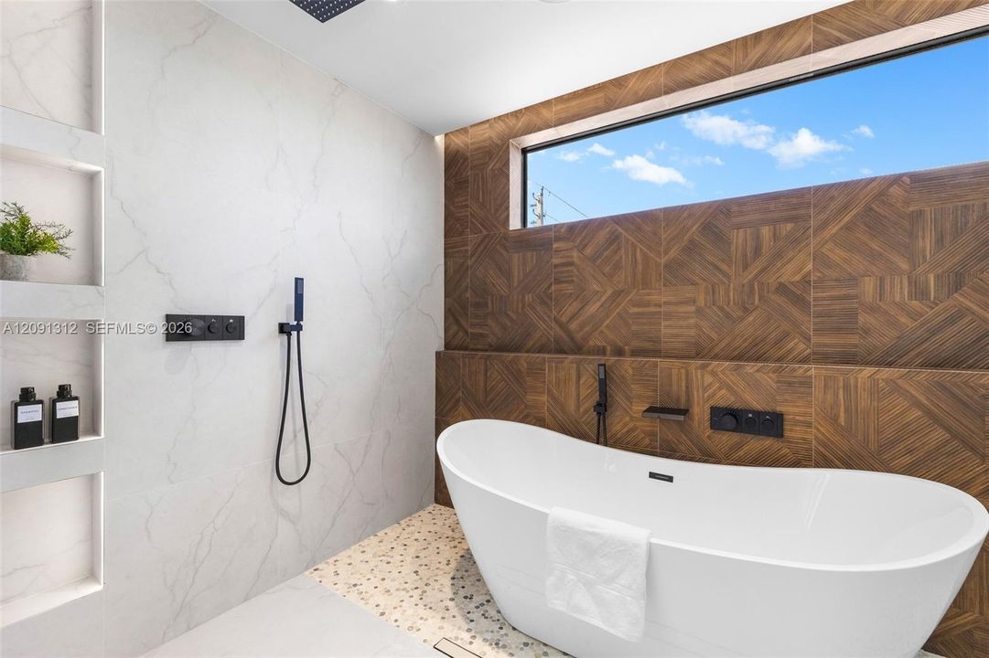Freestanding soaking tub featuring matte black wall-mounted fixtures, geometric wood-patterned accent wall, recessed shelving, and a panoramic clerestory window