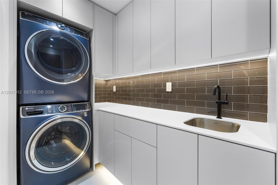 Laundry area featuring stacked front-load appliances, quartz countertops, high-gloss cabinetry, a stainless steel undermount sink with a matte black faucet, and a tiled backsplash