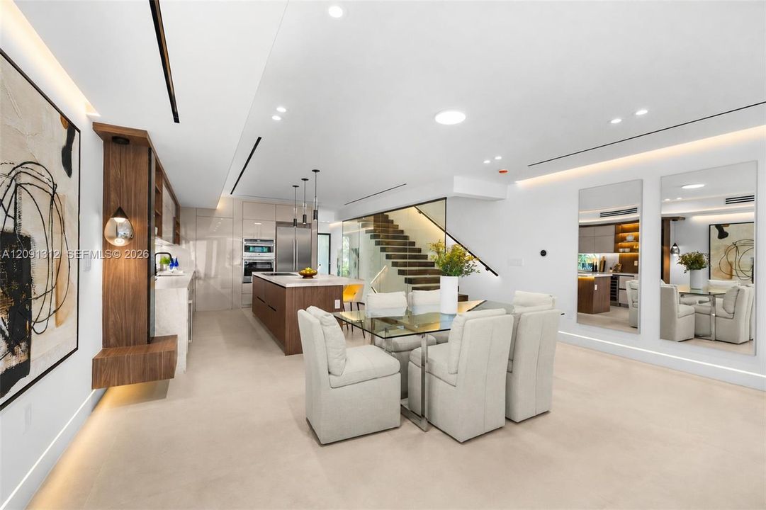 Modern kitchen and dining area featuring high-gloss cabinetry, wood-finish wall paneling with integrated lighting, a stone-top kitchen island, recessed linear ceiling lighting, and a floating wood-tread staircase with a glass railing