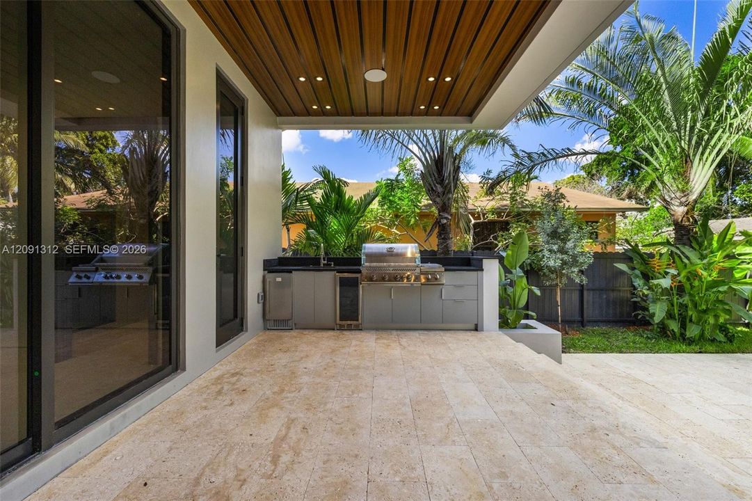 Covered outdoor kitchen featuring built-in cabinetry, stainless steel grill, beverage cooler, stone-finish flooring, and a wood-slat ceiling