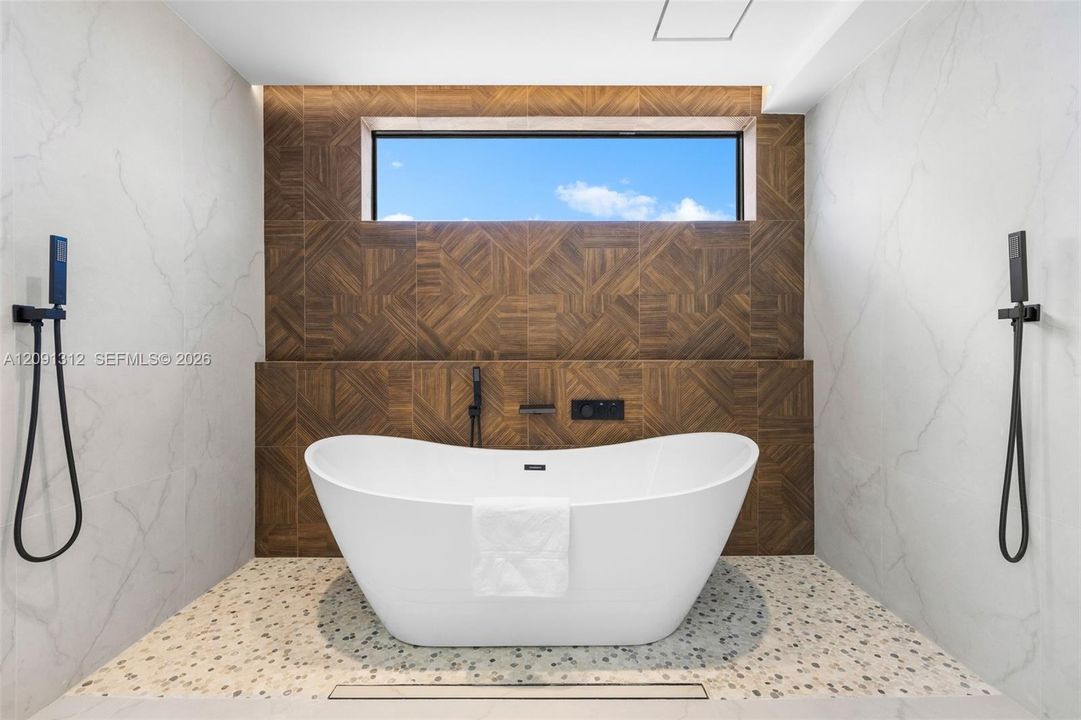 Freestanding soaking tub with matte black handheld shower fixtures, light-toned stone-finish tile surround, geometric wood-patterned accent wall, and horizontal clerestory window