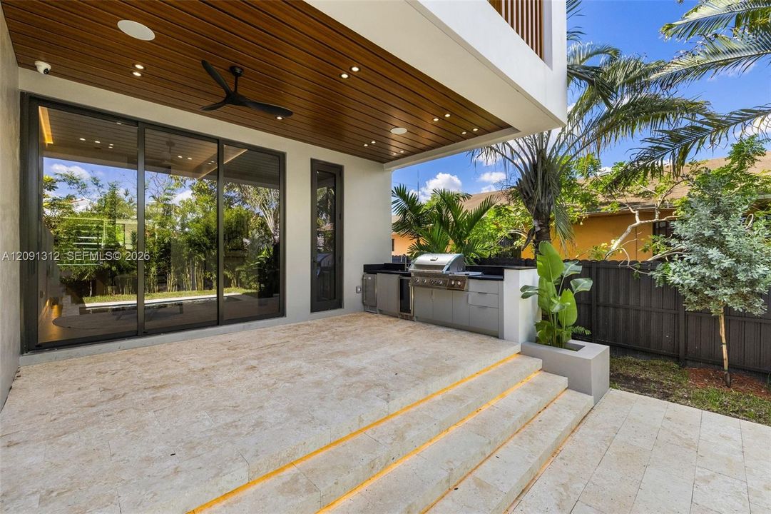 Covered patio featuring a wood-finished tongue-and-groove ceiling with recessed lighting, integrated outdoor kitchen with stainless steel grill, stone-tile flooring, and tiered staircase with step-level lighting