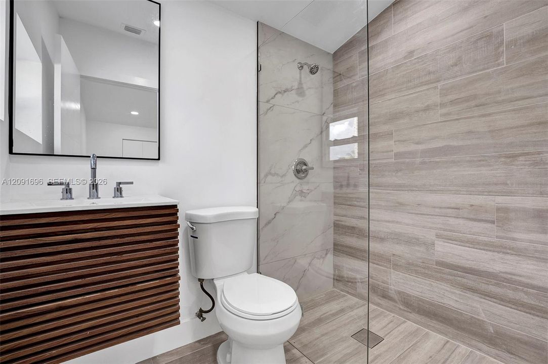 Bathroom featuring a slatted wood-finish floating vanity, rectangular sink with widespread faucet, wall-mounted mirror with a black frame, walk-in shower with a glass panel, and large-format tile surround