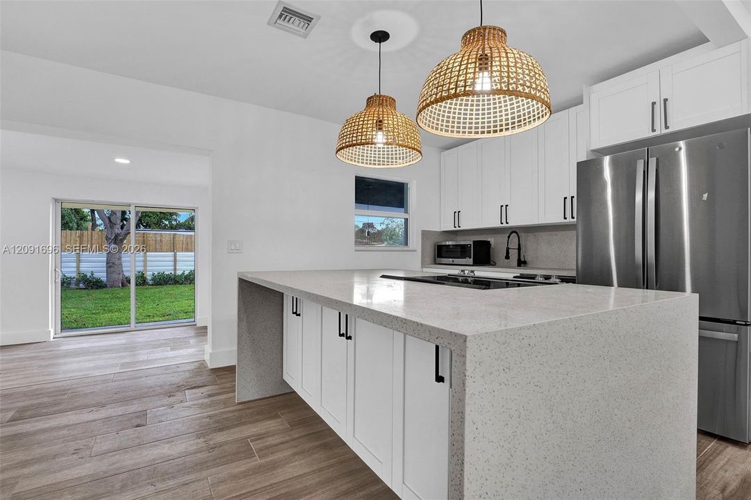 Kitchen featuring a waterfall-edge quartz island, white shaker-style cabinetry, stainless steel refrigerator, textured woven pendant lighting, and wood-finish flooring