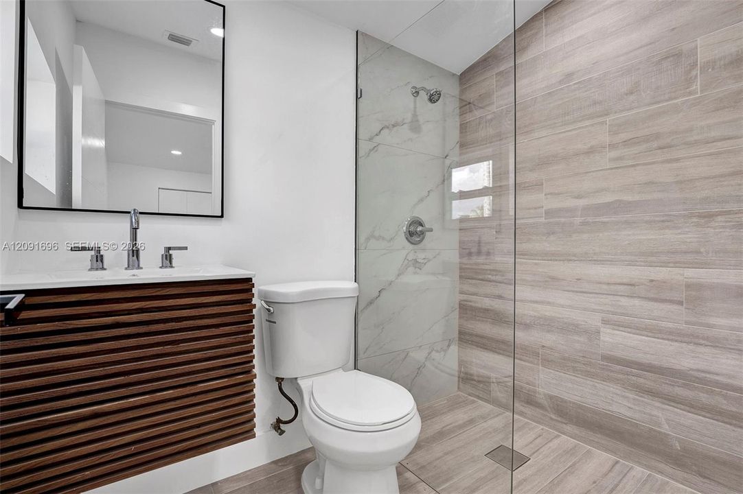 Modern bathroom featuring a wall-mounted slatted wood-finish vanity, large-format tile shower surround with a frameless glass enclosure, and a rectangular mirror with a black frame