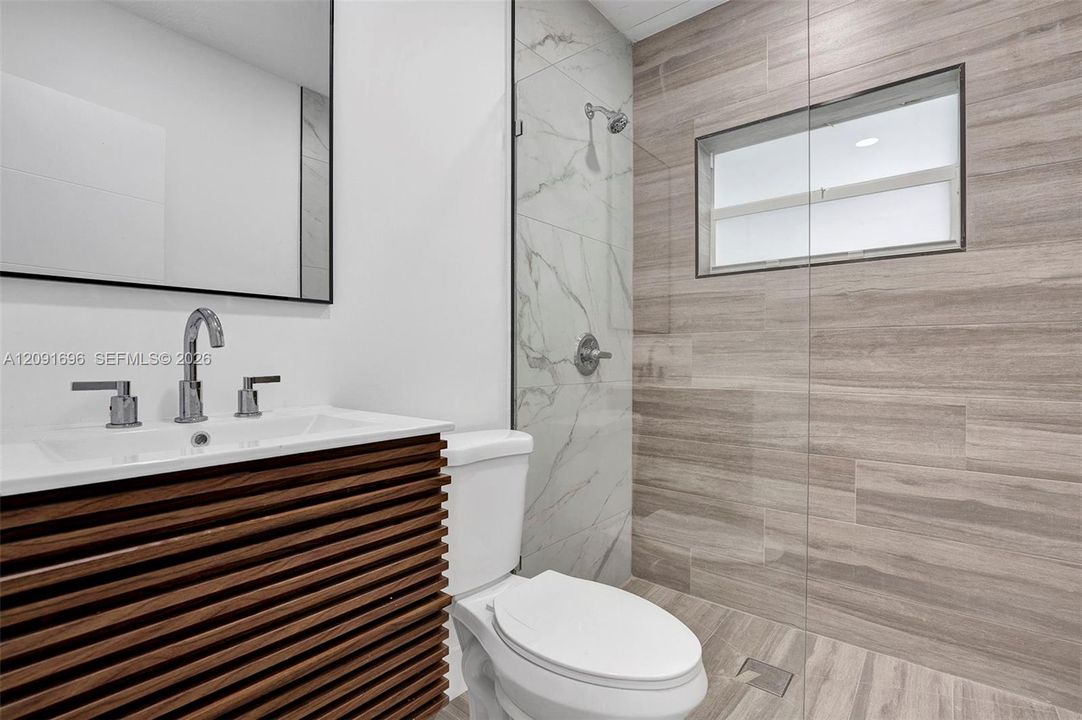 Slatted wood-finish vanity with a rectangular sink, frameless glass walk-in shower featuring dual-finish wall tile, and black-framed mirror
