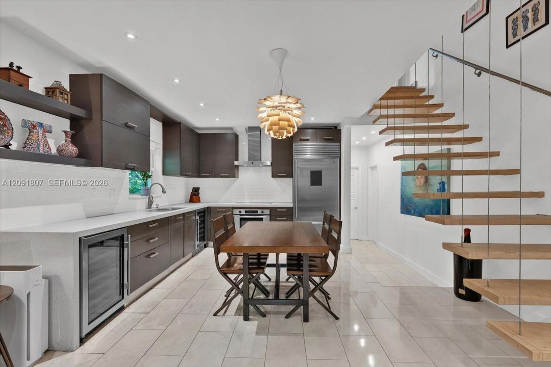 Custom cabinetry with horizontal hardware, light-toned stone tile flooring, stainless steel integrated refrigeration, stone-surfaced countertops, and an open-tread staircase with steel cable railing
