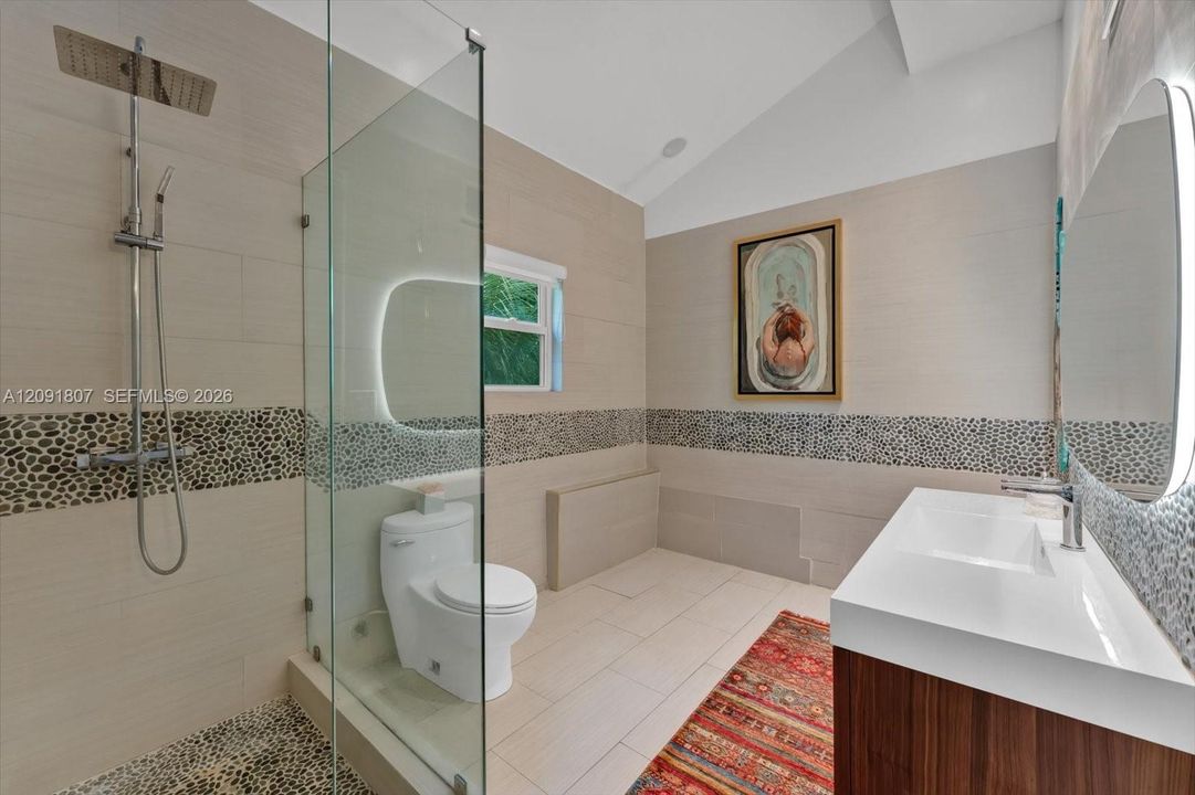Bathroom featuring a glass-enclosed shower with pebble stone flooring and wall accents, a wood-finish vanity with a rectangular sink, wall-mounted illuminated mirrors, and a vaulted ceiling