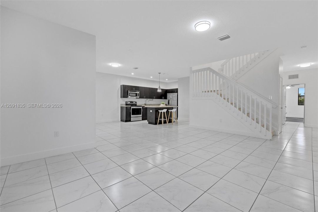 Open-concept interior featuring light-toned tile flooring, kitchen with dark cabinetry, central island, and staircase with white railings