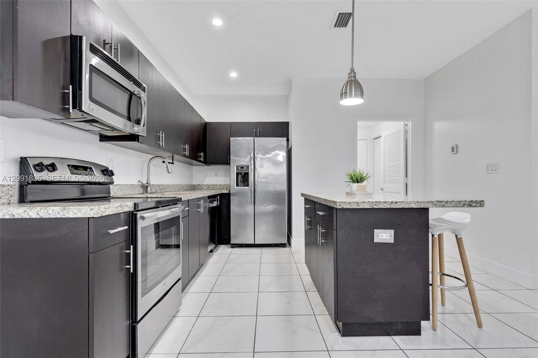 Kitchen featuring flat-panel dark cabinetry, speckled stone-finish countertops, stainless steel appliances, a center island with pendant lighting, and ceramic tile flooring