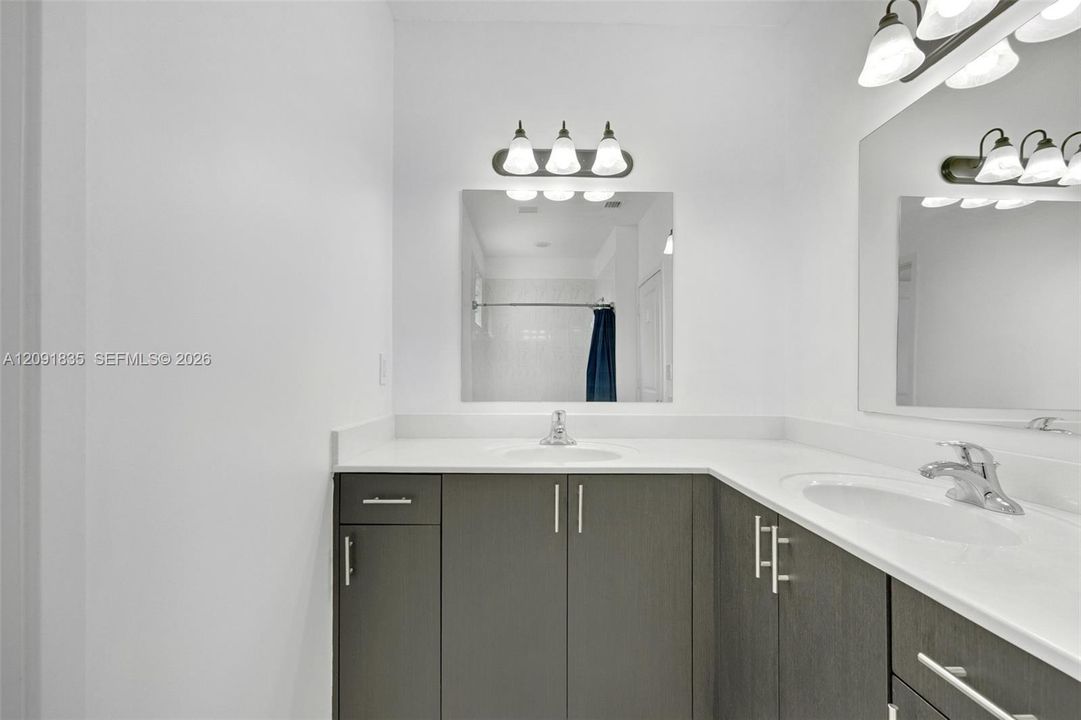 Dual-sink vanity featuring charcoal-toned cabinetry, light-colored solid surface countertops, integrated sinks, polished chrome fixtures, and wall-mounted vanity lighting