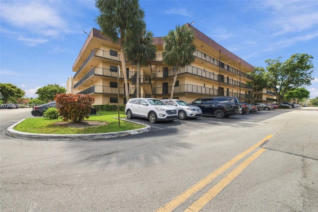 Multi-story residential building exterior featuring private balconies with metal railings, mature palm trees, and asphalt surface parking