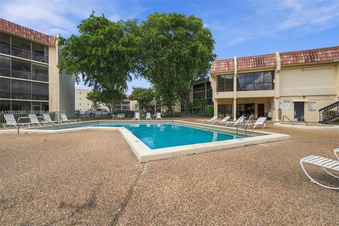 Community swimming pool featuring a textured concrete deck, tile roof architecture, and mature trees