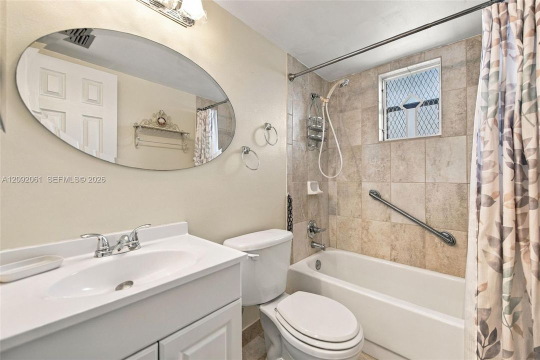 Bathroom featuring a vanity with an integrated basin, an oval wall-mounted mirror, a tiled shower surround, a built-in grab bar, and a window with a lattice security grate