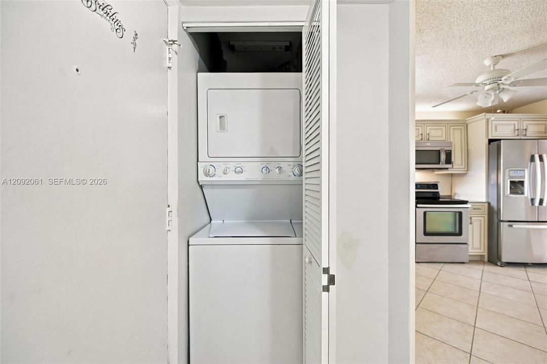 Laundry closet featuring a stackable washer and dryer, louvered bi-fold door, and kitchen area with stainless steel appliances, cabinetry, and tile flooring