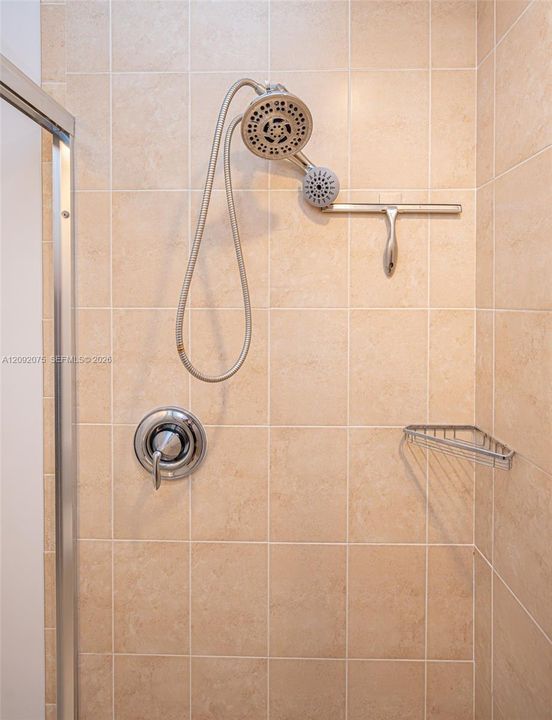 Beige square tile shower surround with a handheld showerhead, chrome fixtures, and a corner shower caddy